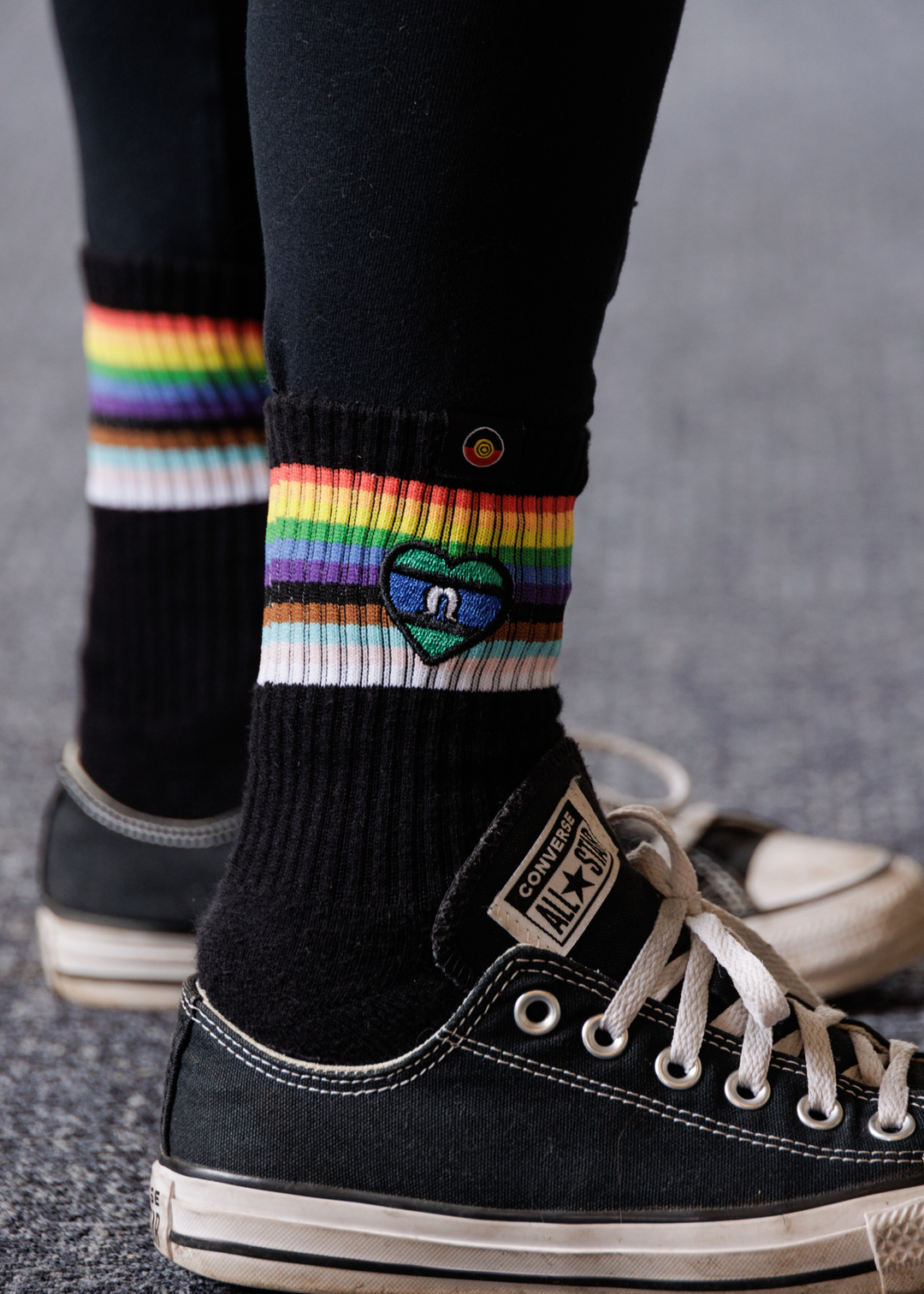 Person wearing black Clothing The Gaps socks with rainbow stripes and a heart design, paired with black sneakers.
