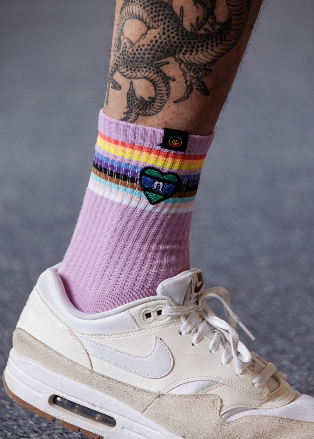 Purple sock with rainbow stripes and a heart design worn with a beige sneaker on a gray background showing TSI flag