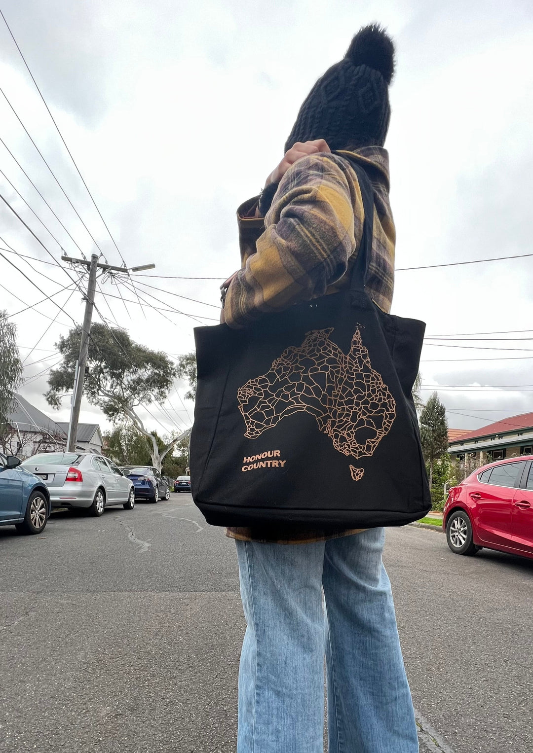 Clothing The Gaps. Decolonise Everyday Bag. Black Tote bag with 2 over the shoulder straps, laptop compartment and small pocket for loose items such as keys inside bag. On outside of bag is a brown screen print featuring the words 'Honour Country' and a large decolonized map of Australia showing all 260 First Nations groups. 