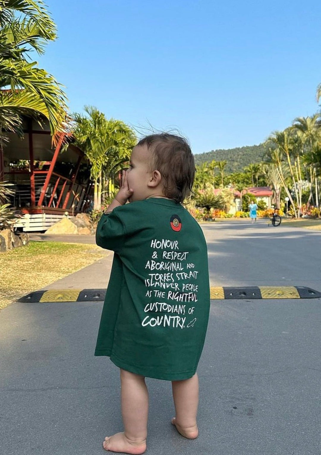 Clothing The Gaps. Kids Handwritten Honour Country Tee. Forrest green T-shirt with 'Honour Country' across chest in handwritten white text and coloured Aboriginal flag small underneath. On Back of tee white hand-writen style text 'Honour and respect Aboriginal and Torres Strait Islander people as the rightful custodians of country.' Circling 'respect' underlining 'custodians' and highlighting 'rightful' in a black colour. Outlined 'bunjil' eagle in bottom right corner.'        