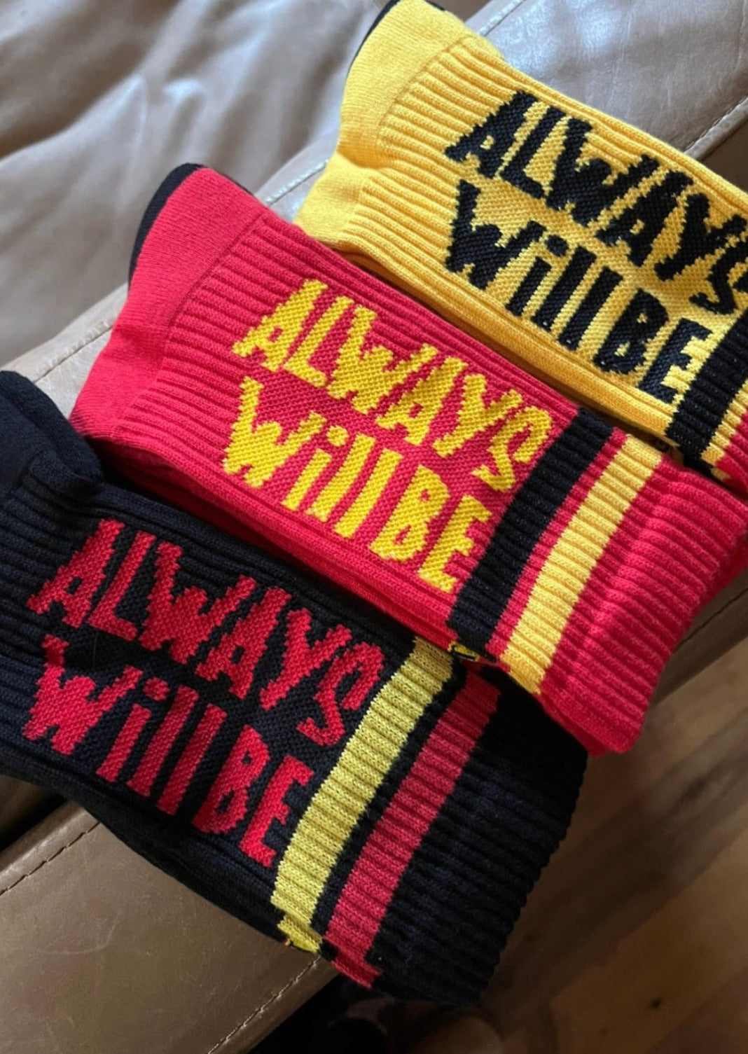 Clothing The Gaps. Power Socks 3 pack. 3 pack red, black and yellow thick cotton socks. Pair 1 Black base sock with 'Always was' on one side of sock and 'Always will be' on other side with red capital text and red and yellow band near top of sock. Pair 2 Red base sock with same text but in yellow and black. With yellow band near top of sock. Pair 3 yellow base sock with same text but in black. With black and red band near top of sock.