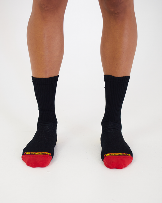 Clothing The Gaps. Long black socks with white Aboriginal flag outline on back. Yellow on heel of socks. Red on toes of socks. Clothing The Gaps written above red section just going over toes.