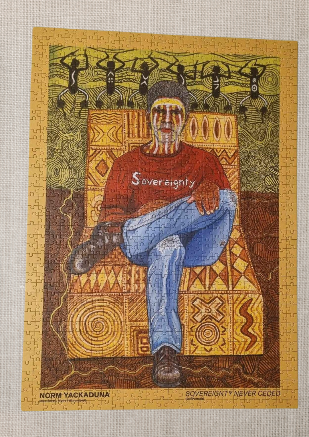 Clothing The Gaps. 1000 piece Puzzle of Aboriginal man with traditional face paint wearing a red sovereignty t-shirt on a yellow possum skin cloak chair. With a brown and green aboriginal style background using dots lines to tell story. Artwork by Norm Yakaduna.