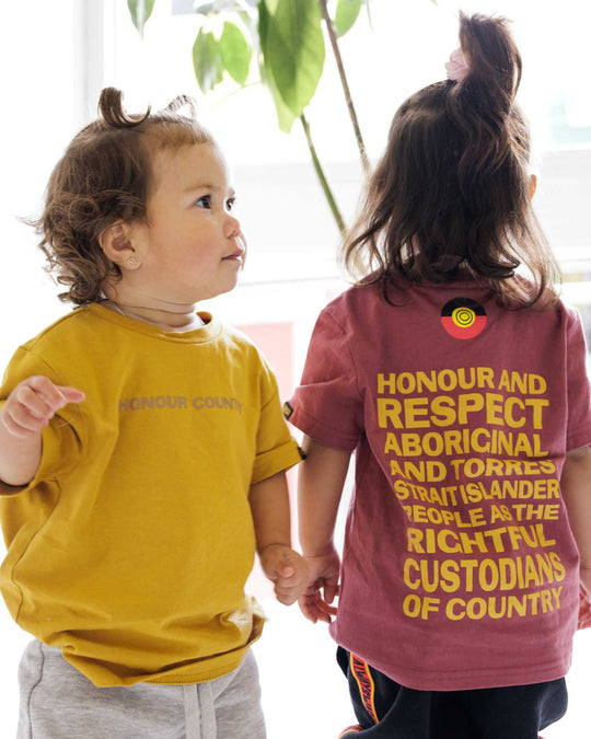 Clothing The Gaps. Kids Mustard Honour Country Tee. Mustard yellow t-shirt with screen printed bold capital text 'Honour and respect Aboriginal and Torres Strait Islander people as the rightful custodians of country.' The word 'Honour country' on the front in the same text and contrasting colour.