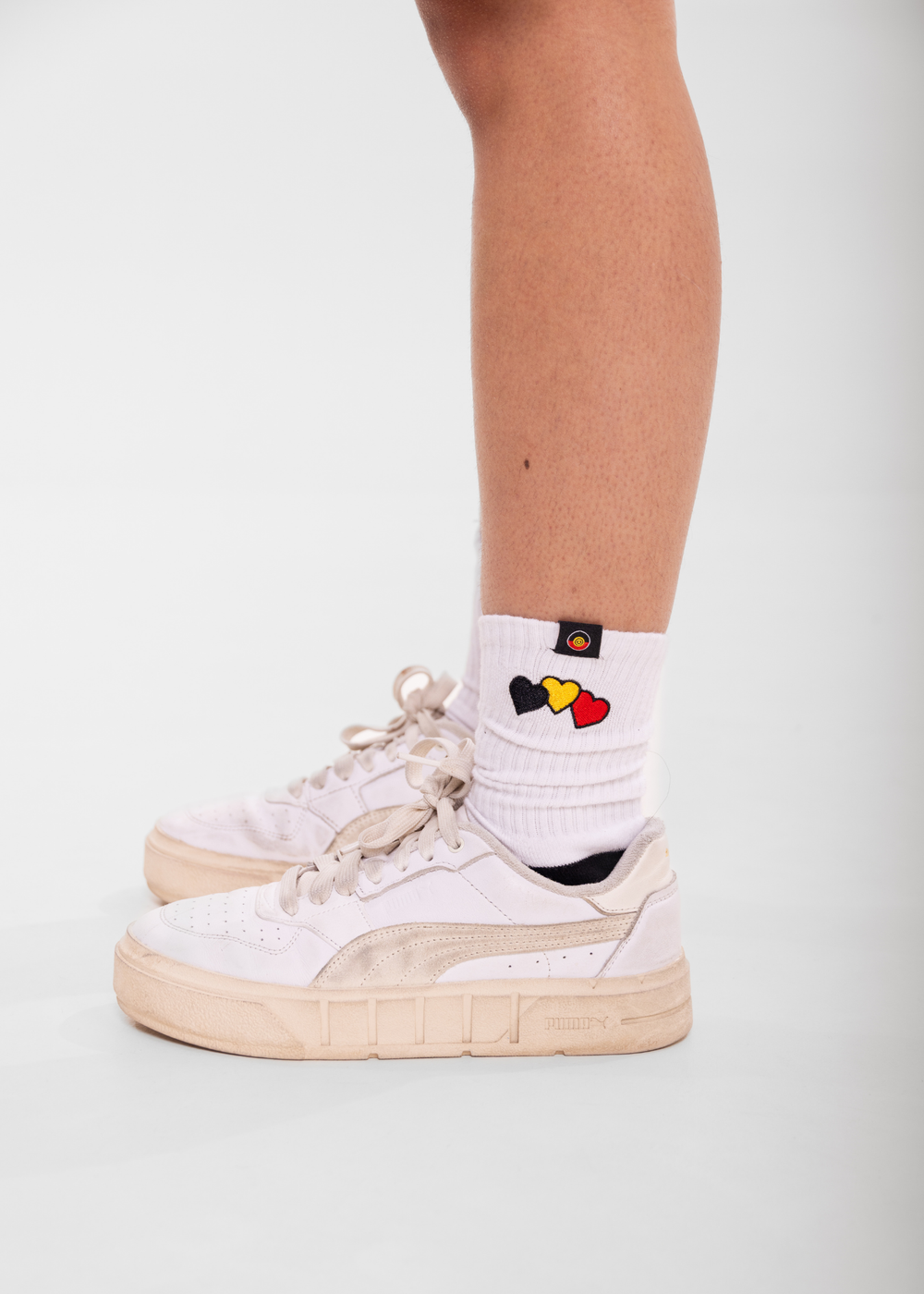 White sneakers with beige soles worn with a white sock featuring colorful heart designs on a light gray background.