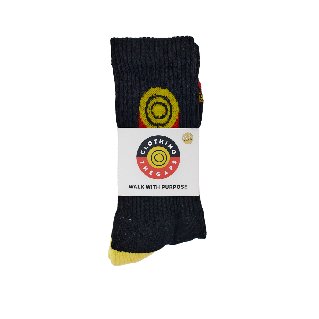 Clothing The Gaps. Flag Rights Socks. Long black socks with rectangle strips going down on both sides of socks representing the Clothing The Gaps logo and Aboriginal flag .With black at the top yellow circle in the middle with 2 black circular rings inside and red underneath the yellow. On both sides of each sock. Yellow on heel of socks. Red on toes of socks. Clothing The Gaps written above red section just going over toes.