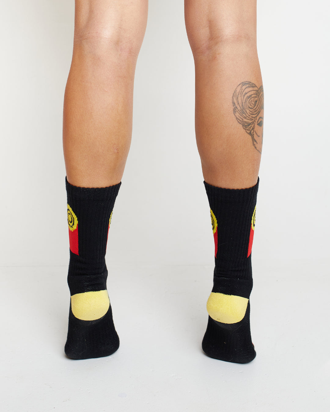 Clothing The Gaps. Flag Rights Socks. Long black socks with rectangle strips going down on both sides of socks representing the Clothing The Gaps logo and Aboriginal flag .With black at the top yellow circle in the middle with 2 black circular rings inside and red underneath the yellow. On both sides of each sock. Yellow on heel of socks. Red on toes of socks. Clothing The Gaps written above red section just going over toes.