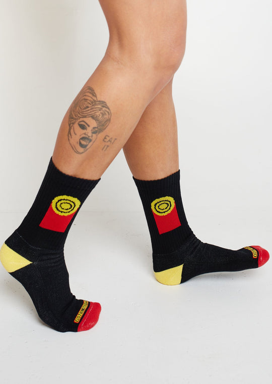 Clothing The Gaps. Flag Rights Socks. Long black socks with rectangle strips going down on both sides of socks representing the Clothing The Gaps logo and Aboriginal flag .With black at the top yellow circle in the middle with 2 black circular rings inside and red underneath the yellow. On both sides of each sock. Yellow on heel of socks. Red on toes of socks. Clothing The Gaps written above red section just going over toes.