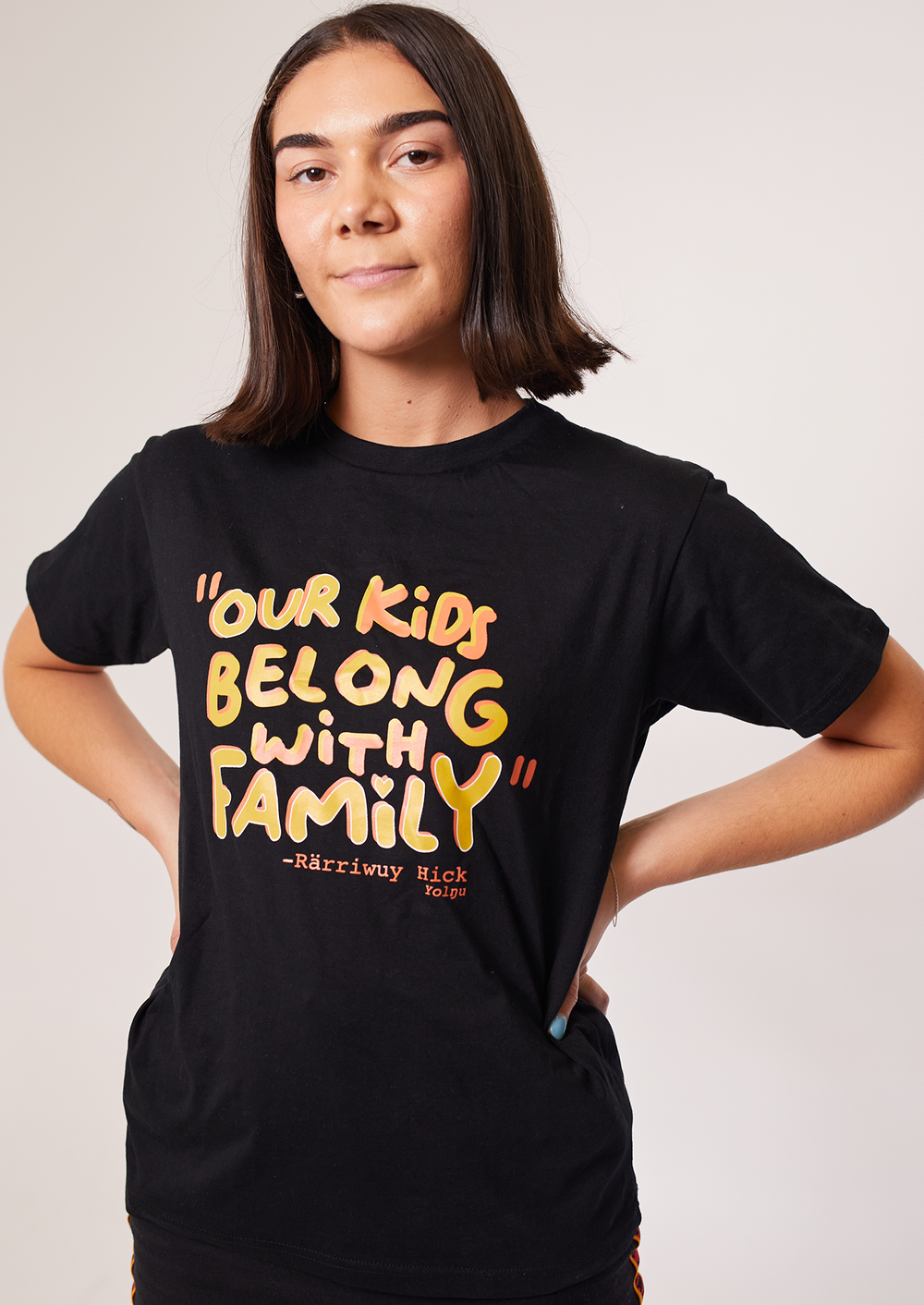 Clothing The Gaps. Our Kids Belong with Family Tee. Black T-shirt with the quote “Our kids belong with family” Rarriwuy Hick Big on front chest of t-shirt in yellow orange text.