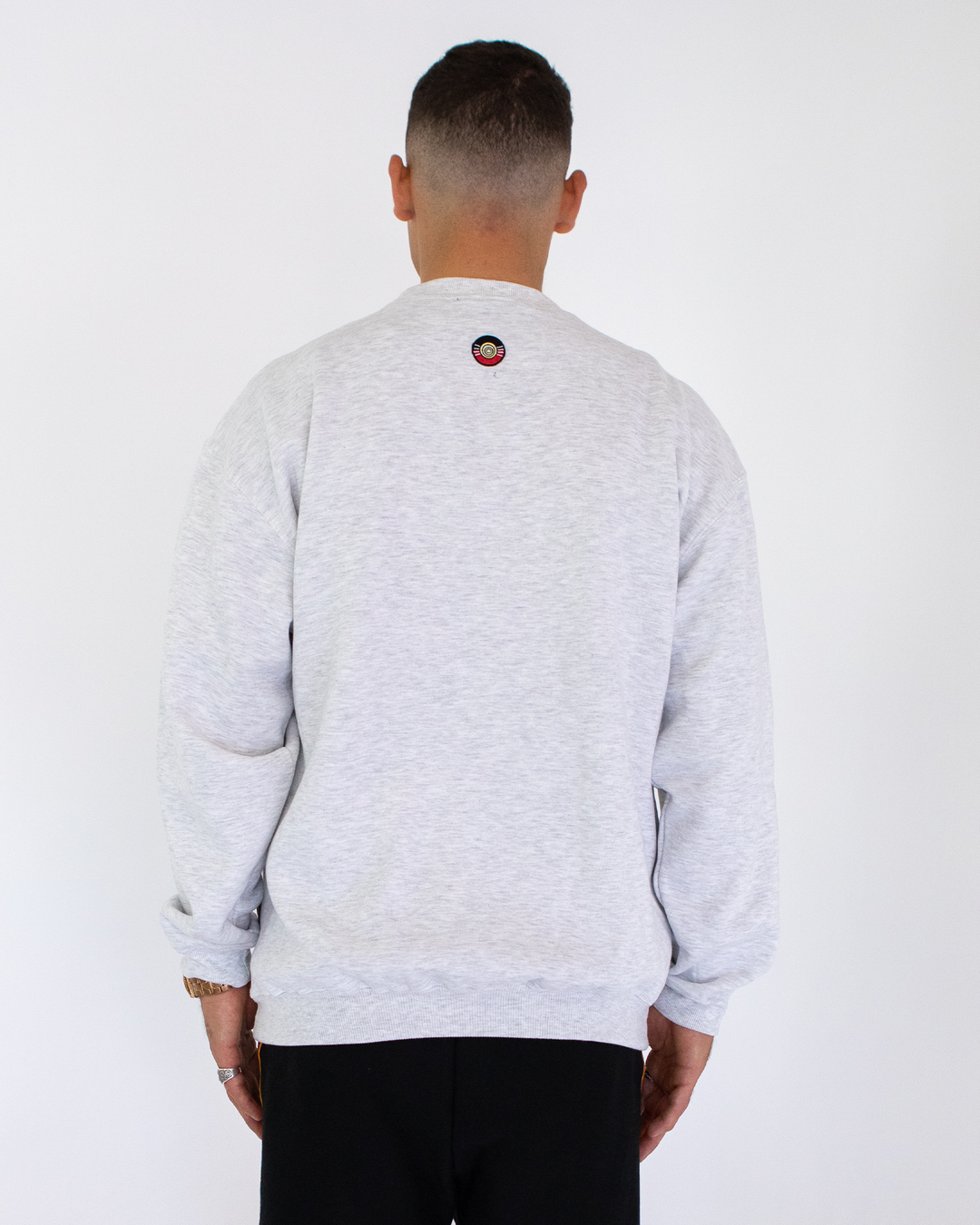 Clothing The Gaps. Classic Crew Jumper. This light grey crew jumper features a minimalist front embroidery piece proudly showcasing the words "Clothing The Gaps" in black with a yellow and red outline. Underneath a white embroidered outline in white of the Aboriginal flag.