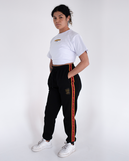 Clothing the Gaps. Black cotton Pants. With black drawstrings. Elastic cuts and waistband. Embroiled Clothing The Gaps logo. Black 'always was always will be' text on a red background with a yellow trim. In a cotton tape strip going down both sides of pants.