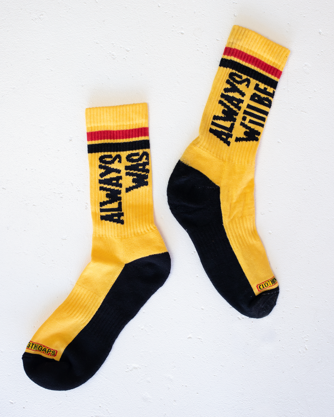 Clothing The Gaps. Power Socks 3 pack. 3 pack red, black and yellow thick cotton socks. Pair 1 Black base sock with 'Always was' on one side of sock and 'Always will be' on other side with red capital text and red and yellow band near top of sock. Pair 2 Red base sock with same text but in yellow and black. With yellow band near top of sock. Pair 3 yellow base sock with same text but in black. With black and red band near top of sock.