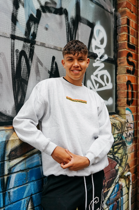 Clothing The Gaps. Classic Crew Jumper. This light grey crew jumper features a minimalist front embroidery piece proudly showcasing the words "Clothing The Gaps" in black with a yellow and red outline. Underneath a white embroidered outline in white of the Aboriginal flag.