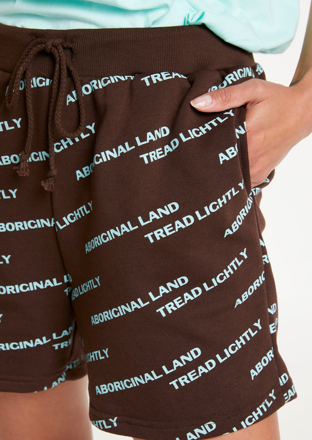 Clothing The Gaps. Blue Tread Lightly shorts. Brown shorts with repeating pattern all over crew of the words 'Aboriginal Land Tread Lightly' text in a light blue colour. Includes solid brown elastic waist with brown drawstrings and has 2 deep pockets on both sides.