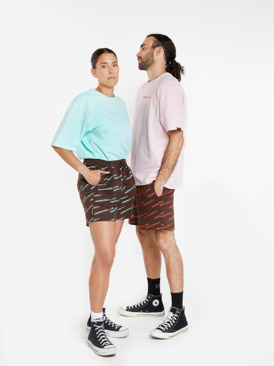 Clothing The Gaps. Orange Tread Lightly shorts. Brown shorts with repeating pattern all over crew of the words 'Aboriginal Land Tread Lightly' text in a orange colour. Includes solid brown elastic waist with brown drawstrings and has 2 deep pockets on both sides.